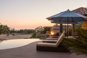 Pool lounge chairs with umbrellas and outdoor firepit at Inspirato Los Cabos villa at sunset.