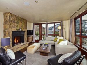 Living room at Solaris Vail residence with beige couch, two arm chairs, stone fireplace, and mountain views.