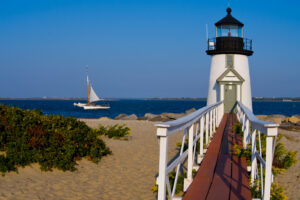 White and black lighthouse with red ramp on beach shore with white sailboat in Cape Cod, Massachusetts.