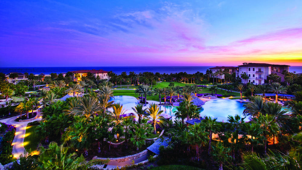 Pool area of Esperanza hotel with manicured gardens and palm trees with the ocean in the background during a sunset.