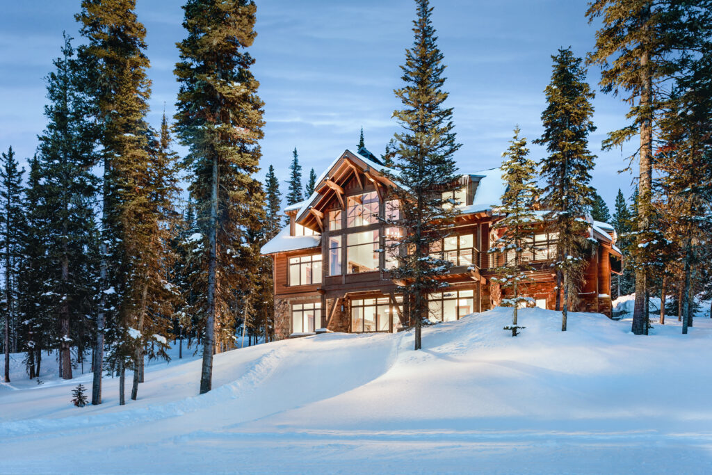 An Inspirato luxury mountain home in Big Sky, Montana lit up at night, tucked within the forest with snow during winter.
