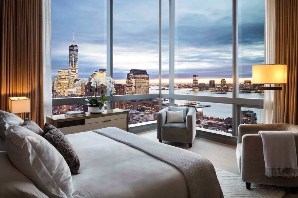 SoHo at The Dominick hotel bedroom with king bed with floor to ceiling windows with views of the New York City skyline at night with the light illuminated. 