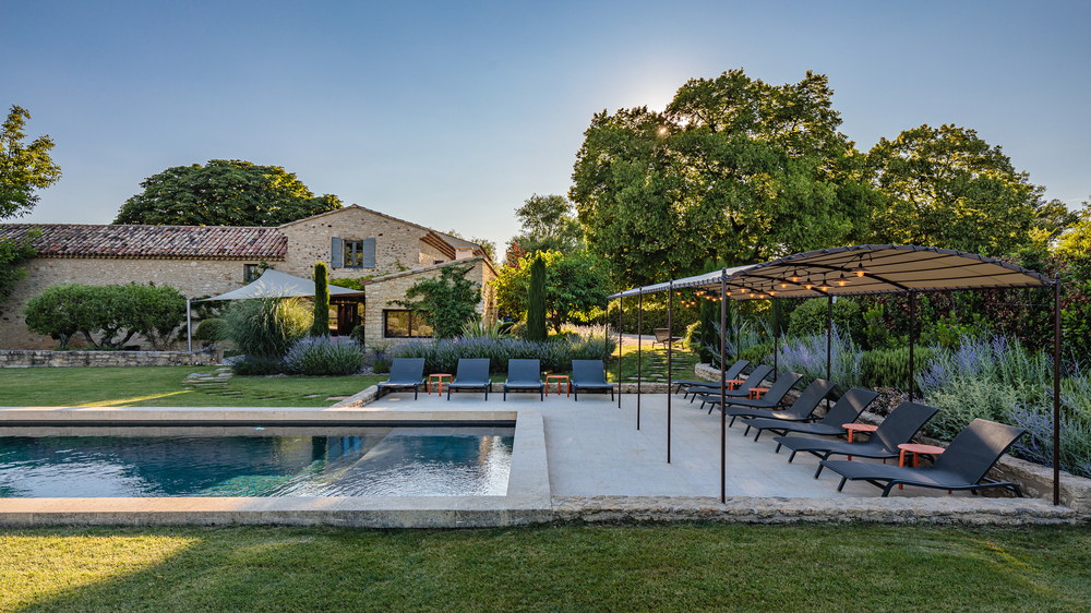 Pool area of Maison du Calavon pool area with rectangle pool with lounge chairs, some under a covering and the rear exterior of the stone home.