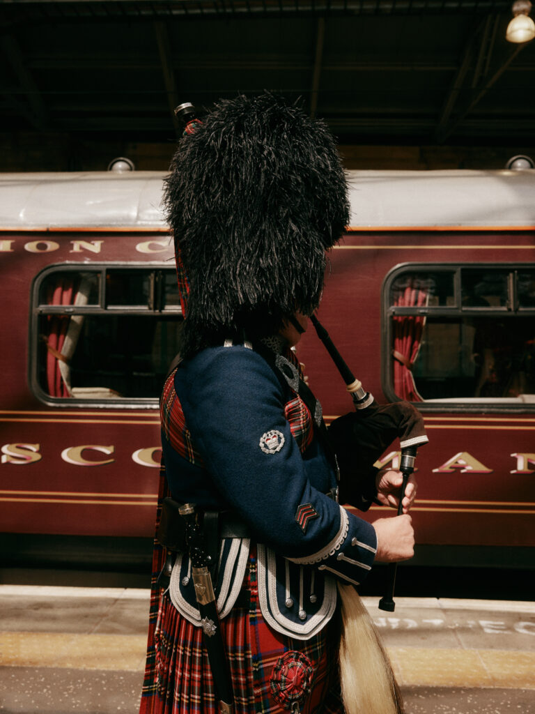 A traditional Scottish bagpiper in full regalia stands beside the Belmond Royal Scotsman luxury train.