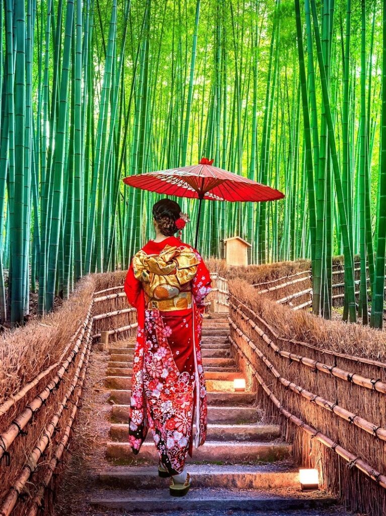 A woman in a vibrant red kimono and traditional umbrella walks through the serene Arashiyama Bamboo Grove in Kyoto, Japan.
