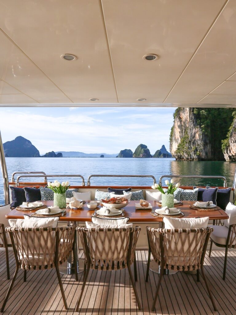 An elegant alfresco dining setup on a private yacht with views of Thailand’s limestone cliffs and tranquil blue waters.