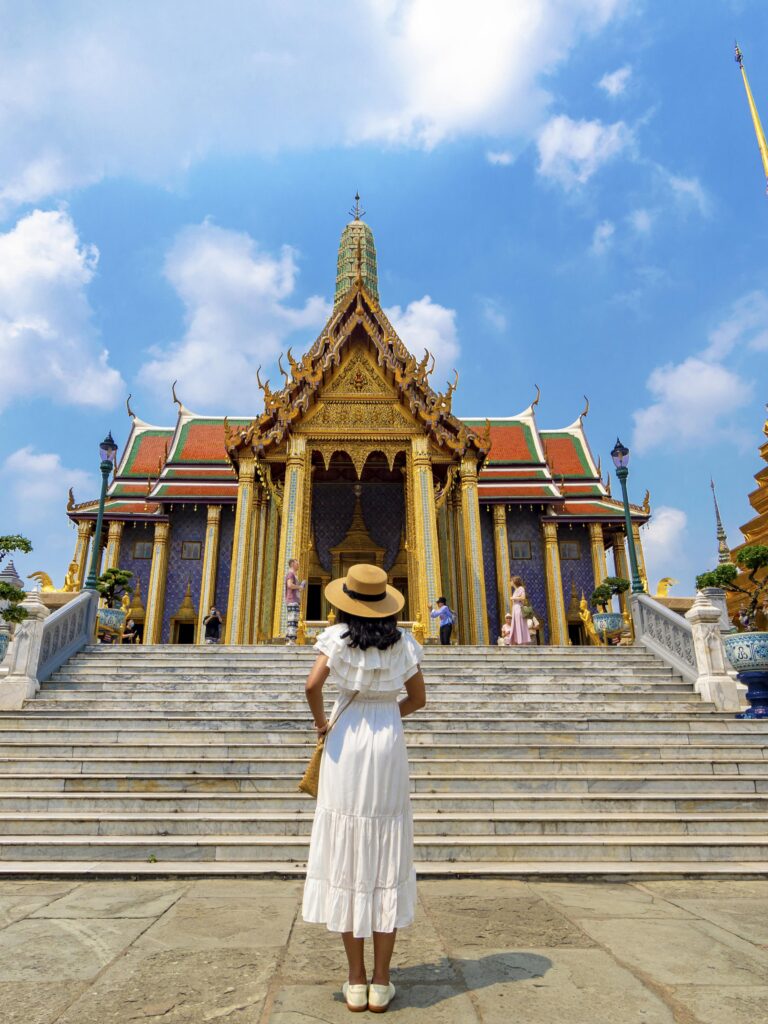 A woman in a white dress stands before the ornate entrance of Wat Phra Kaew, the Temple of the Emerald Buddha in Bangkok.