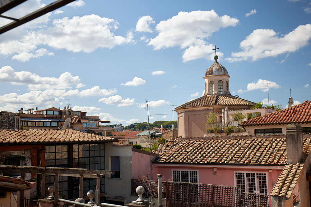 Scenic rooftop view over Rome with terracotta-tiled buildings and a historic domed chapel under a bright blue sky with fluffy clouds.