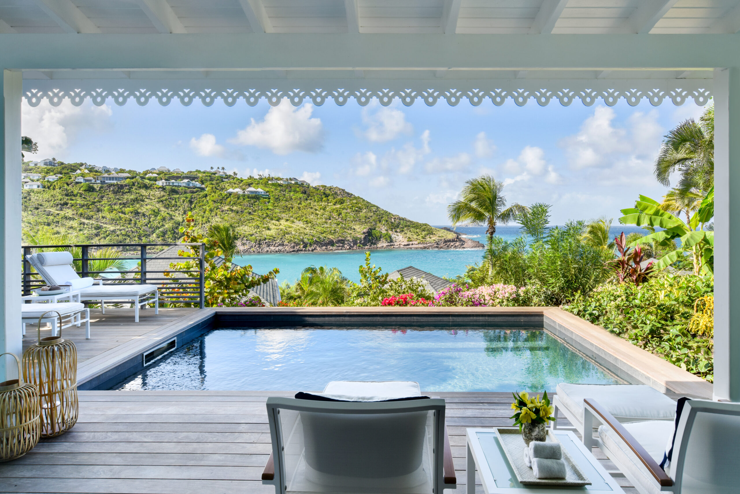 Luxurious tropical villa terrace with a private infinity pool overlooking turquoise ocean waters and lush green hills, surrounded by vibrant flowers and palm trees under a bright blue sky.