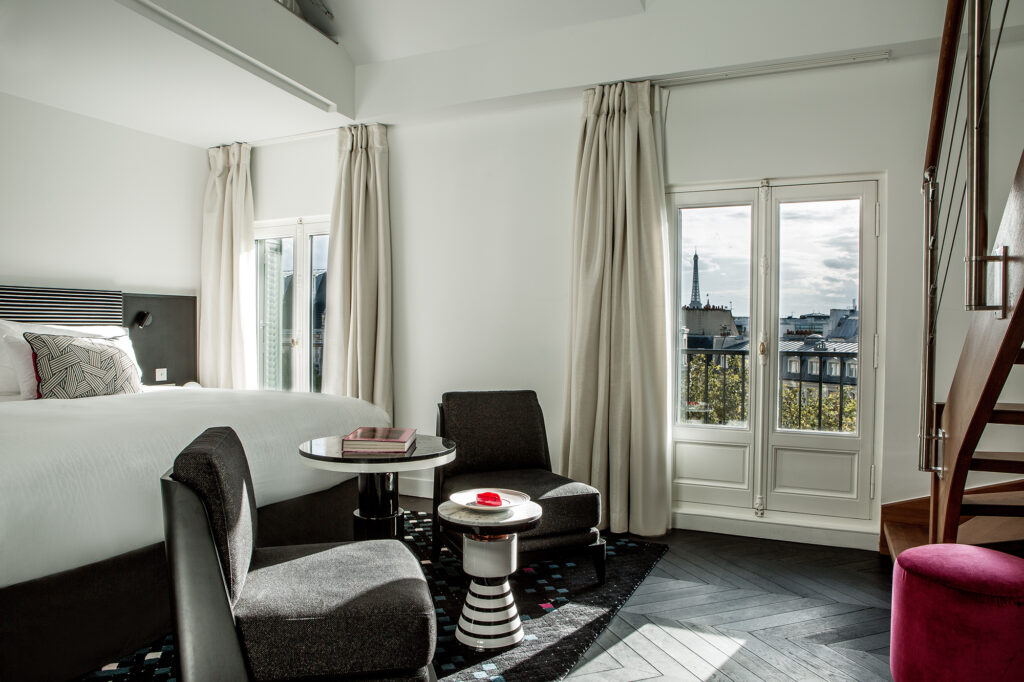 Elegant modern hotel room with a large bed, cozy seating area, and staircase, featuring floor-to-ceiling windows that offer a view of Paris rooftops and the Eiffel Tower in the distance.