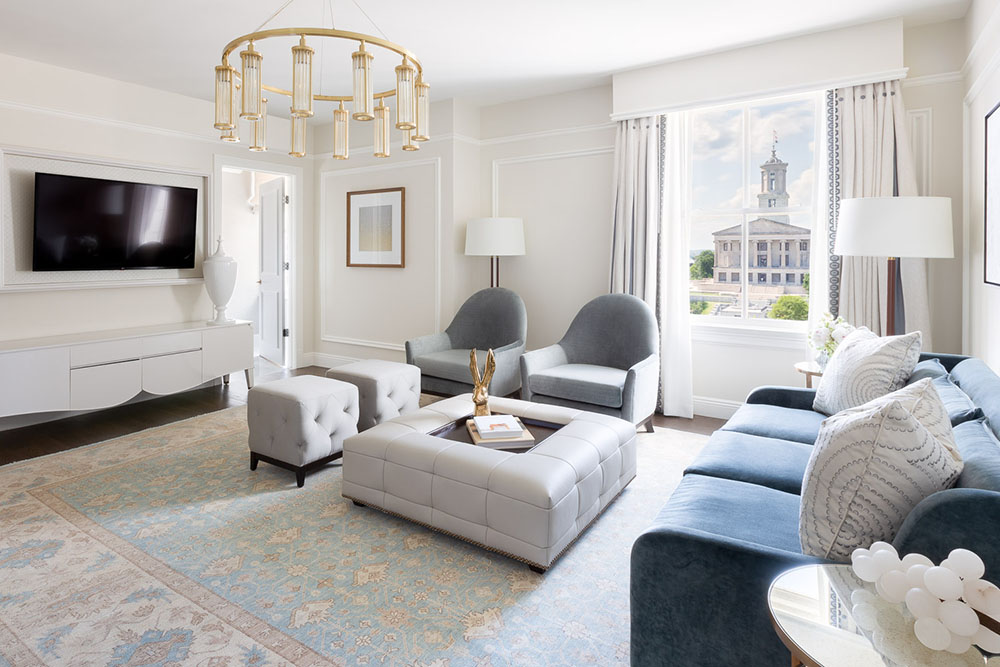 Elegant executive suite at The Hermitage Hotel in Nashville featuring a plush blue sofa, tufted ottoman, modern chandelier, and views of the Tennessee State Capitol.