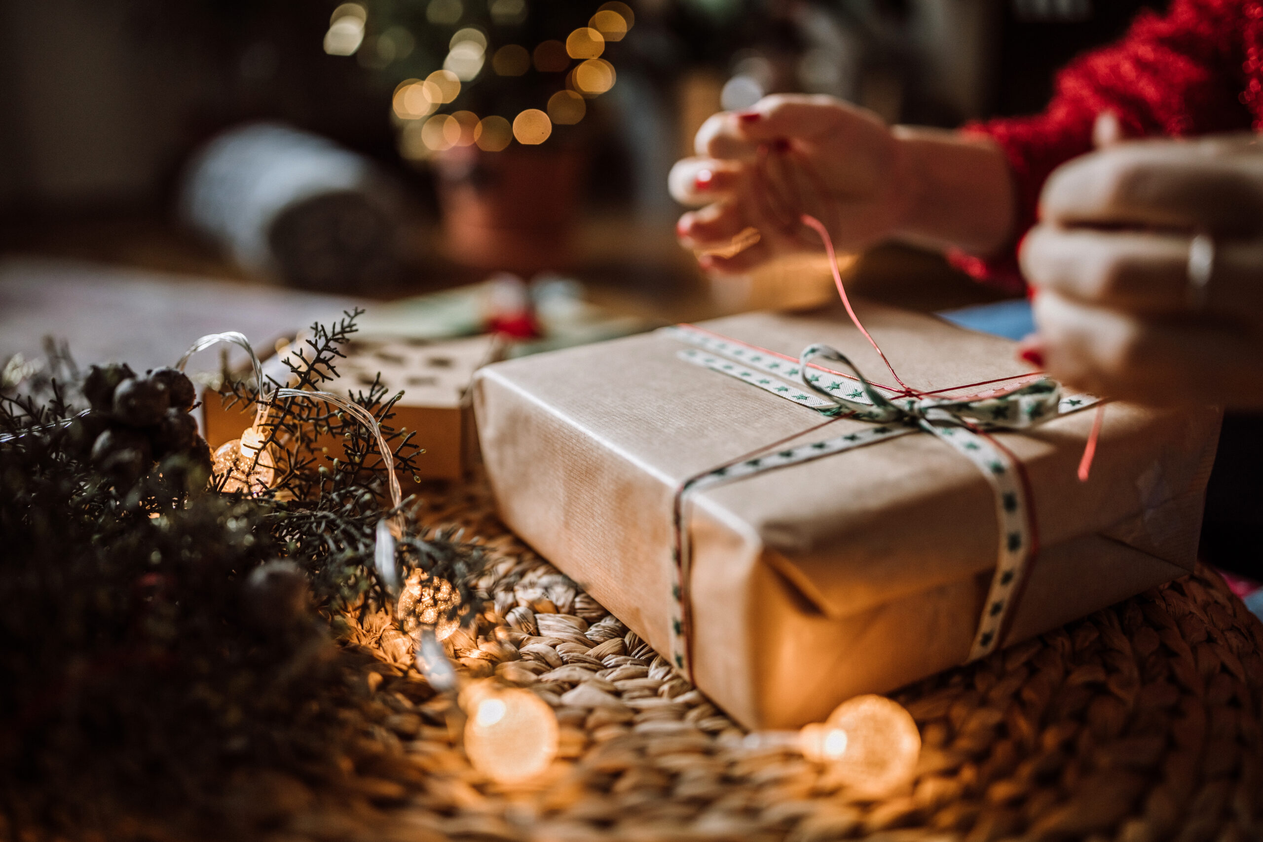 Close-up of hands tying ribbon on a holiday gift wrapped in brown paper, surrounded by warm string lights, evergreen sprigs, and festive décor—capturing the cozy, personal joy of holiday gift-giving.