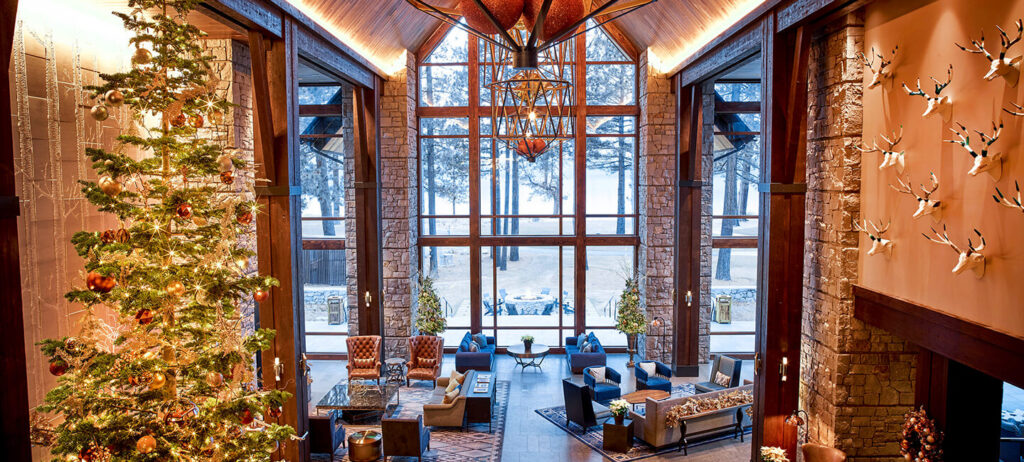 Festive lodge interior with a tall, sparkling Christmas tree, rustic stone walls, and floor-to-ceiling windows overlooking snowy pines—perfect for a Hallmark-style luxury holiday escape