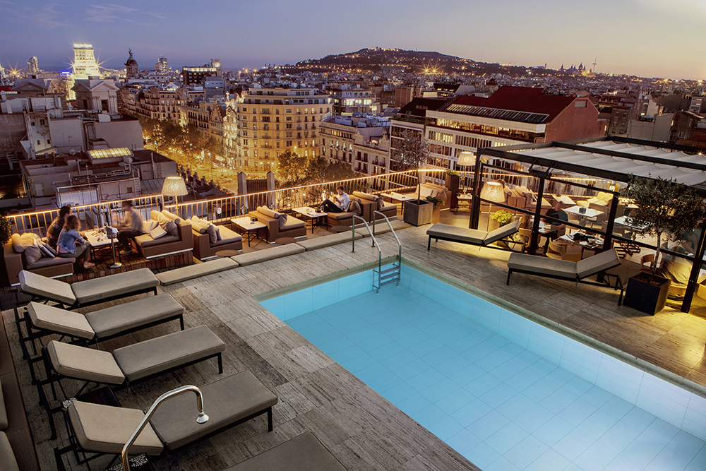Rooftop pool and terrace at a luxury hotel in Barcelona, Spain, overlooking the illuminated cityscape at dusk with lounge seating and scenic views of Montjuïc.