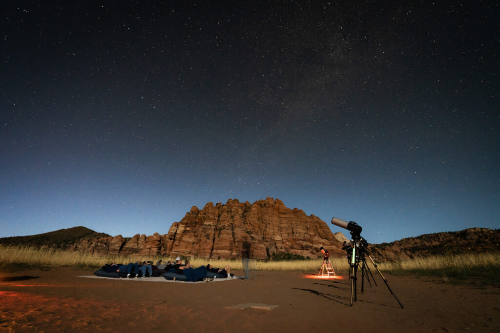 Guests stargaze on a desert floor beneath a clear night sky in Southern Utah, with telescopes set up and red rock formations silhouetted in the background.