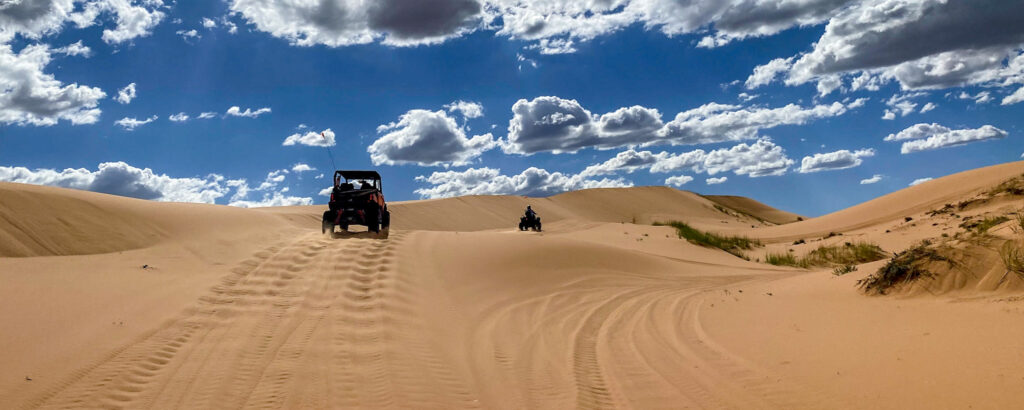 Guests drive UTVs across rolling sand dunes under a bright blue sky with scattered clouds during an off-road adventure in Southern Utah.