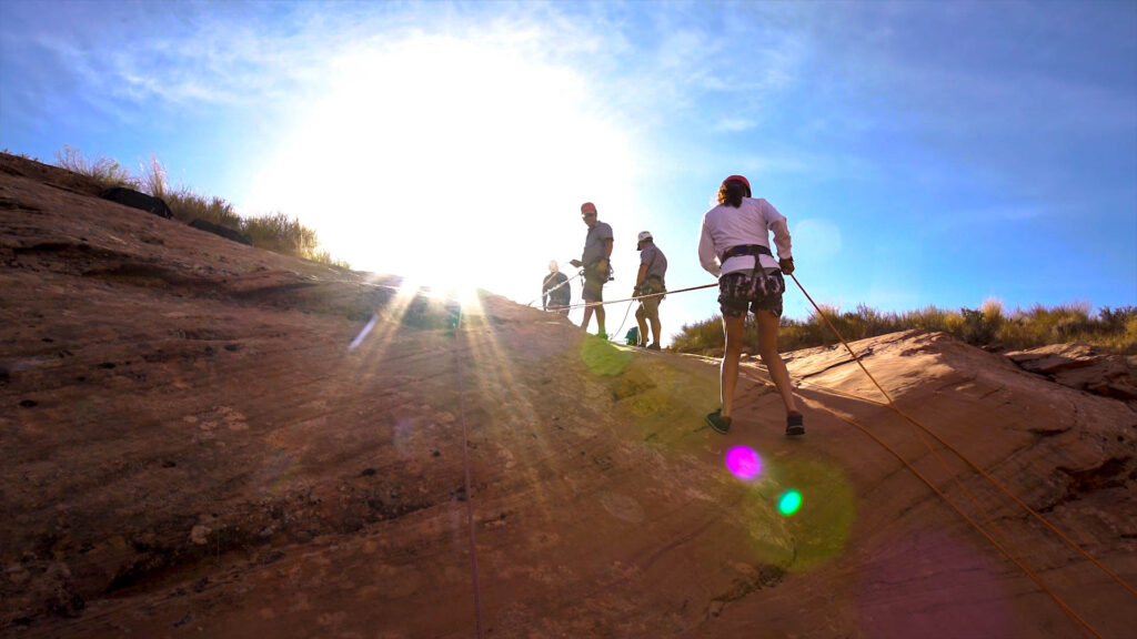 Guests participate in a guided canyoneering experience on sunlit red rock terrain in Southern Utah, wearing helmets and harnesses under a clear blue sky.