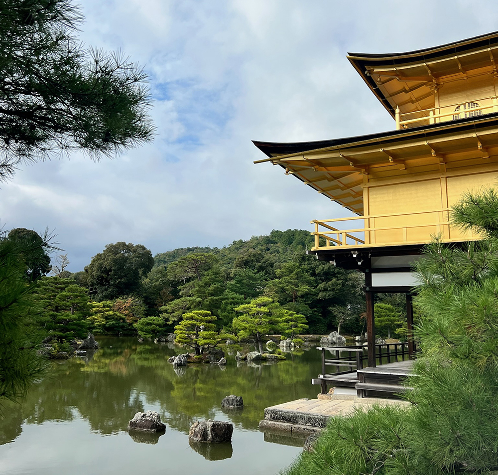 Side view of Kinkaku-ji, the Golden Pavilion in Kyoto, Japan, reflected in a tranquil pond and surrounded by lush pine trees and traditional gardens under a cloudy sky.
