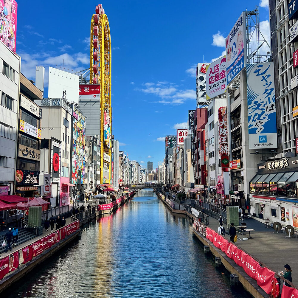 Daytime view of Dotonbori Canal in Osaka, Japan, lined with colorful billboards and restaurants, with the iconic yellow Don Quijote Ferris Wheel towering above the vibrant street scene.
