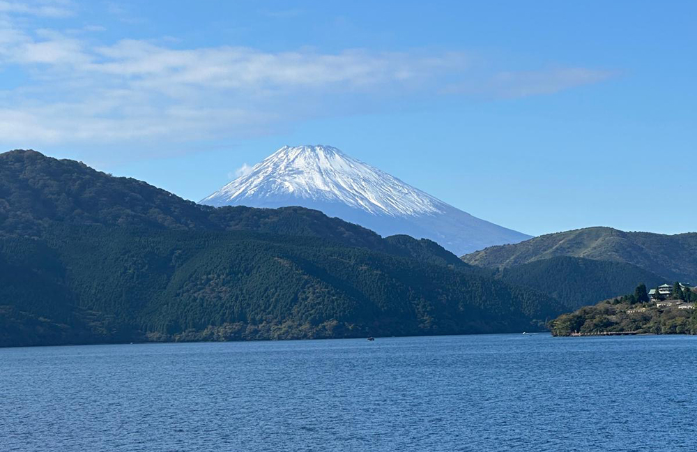 Snow-capped Mount Fuji rising above the forested hills of Hakone, Japan, with Lake Ashi in the foreground under a clear blue sky.