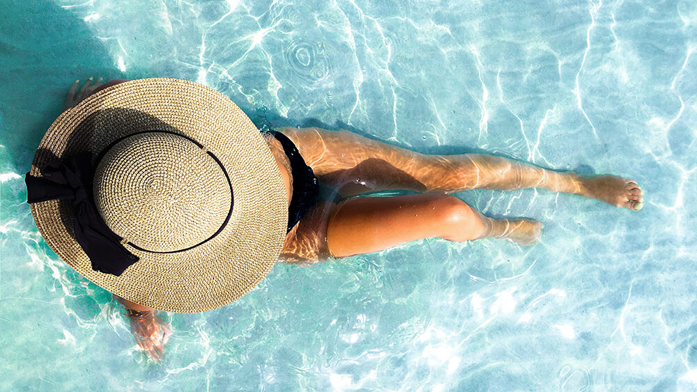 Woman relaxing in a sunlit pool wearing a wide-brimmed hat—capturing the essence of a serene, luxury beach getaway with Inspirato