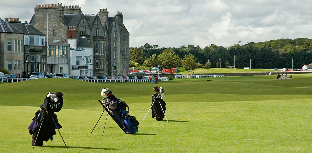 Golf bags on the Old Course at St. Andrews in Scotland with historic clubhouse and green fairways — an Inspirato luxury golf vacation destination for him.