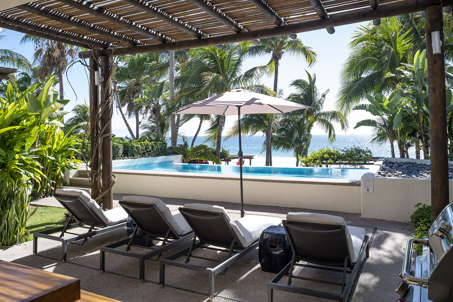 Luxurious beachfront patio with lounge chairs facing a private infinity pool, framed by tropical palm trees and overlooking a serene ocean view under a clear blue sky.