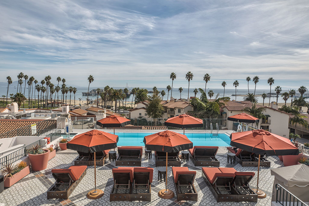 Rooftop pool with red loungers and umbrellas overlooking the Pacific Ocean and palm-lined beach—luxury coastal retreat in Santa Barbara with Inspirato