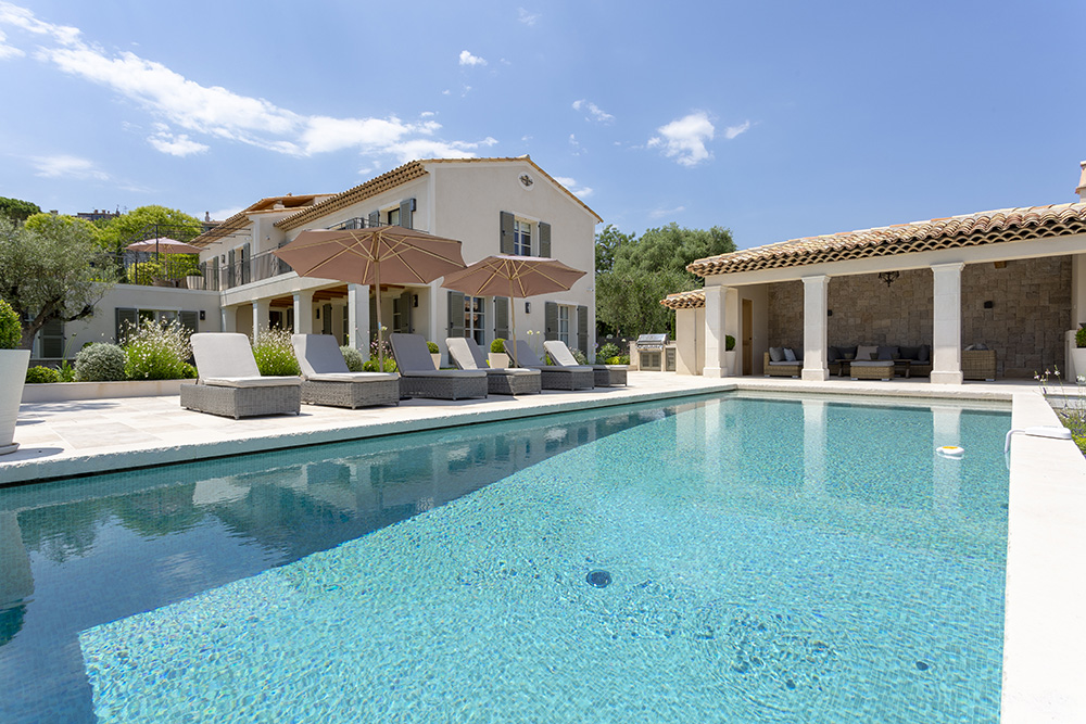 Luxury Mediterranean villa with terracotta roof tiles and light-colored stucco walls, surrounded by lush landscaping. A sparkling turquoise swimming pool is in the foreground, bordered by cushioned lounge chairs and large beige umbrellas. An outdoor covered seating area with stone walls completes the serene, sun-soaked setting.