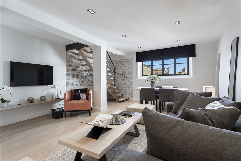 Modern living room with minimalist décor, featuring a cozy seating area with gray sofas, a wooden coffee table, and a coral accent chair. A flat-screen TV is mounted on the wall above a floating shelf, and a stone accent wall with an open staircase adds rustic charm. A window-lit dining area sits at the back with a black-out shade and neutral-toned furniture.