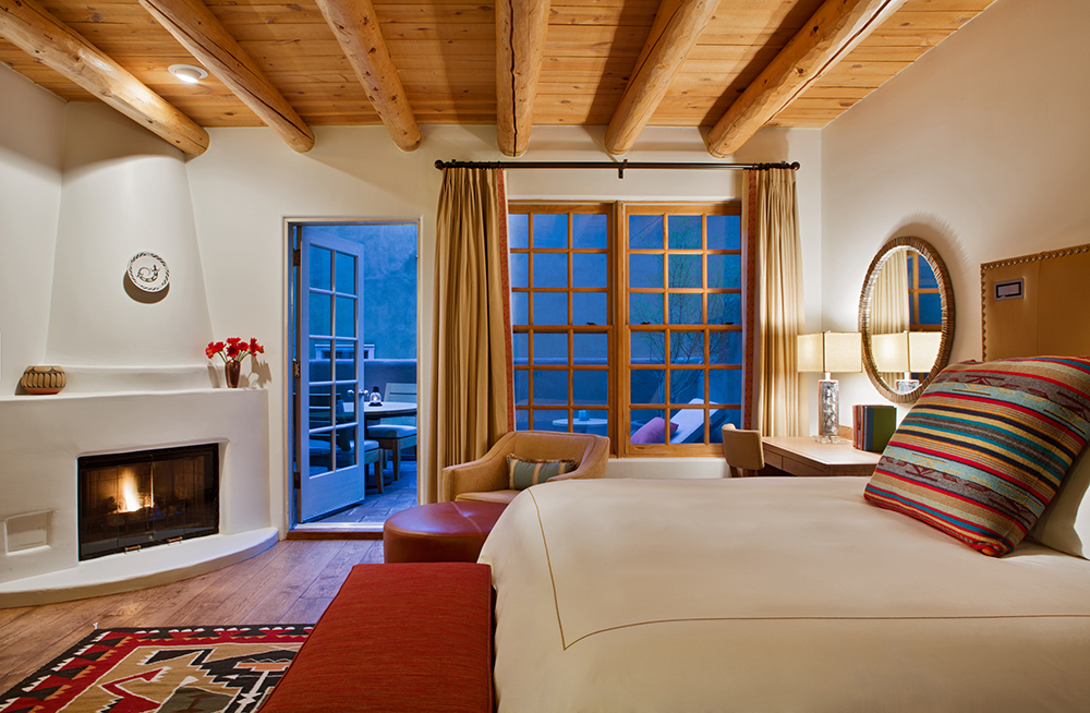 A cozy Southwestern-style hotel room features a wooden beamed ceiling, a corner fireplace, and a plush bed with a colorful striped pillow. The room opens onto a private balcony through French doors, revealing outdoor seating. Warm lighting and earthy tones create a welcoming, intimate ambiance.