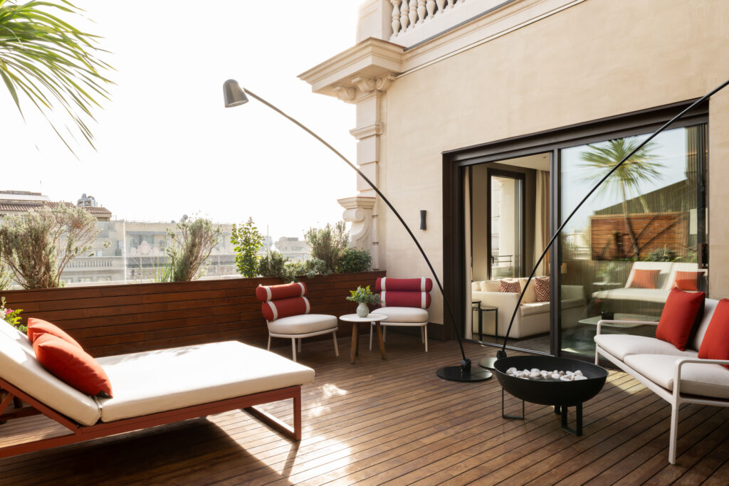 A stylish private terrace of the Almanac Barcelona Terrace Suite, featuring a warm wood deck with modern outdoor furniture. A cushioned lounge chair with a red pillow is positioned for sunbathing, while a cozy seating area with red and white chairs surrounds a small table. A sleek black fire pit and sculptural floor lamp add a contemporary touch. Potted plants line the edge, offering a bit of privacy and greenery, and sliding glass doors open into a bright and elegant interior living space.