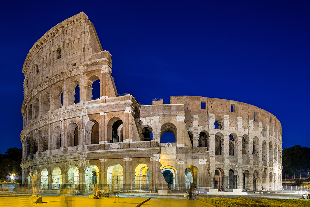 The Colosseum in Rome illuminated at night, showcasing its iconic ancient Roman architecture with arched walls, weathered stone, and a deep blue evening sky in the background. A few blurred figures add motion to the historic setting.