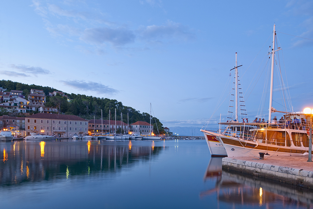 A tranquil harbor at dusk in a coastal Croatian town. The calm water reflects the soft glow of lamplights and the silhouettes of sailboats and yachts docked along the waterfront. A large wooden boat with passengers aboard is moored in the foreground on the right. Stone buildings with red-tiled roofs and hillside homes are nestled among trees, while the sky transitions into soft shades of blue with a few scattered clouds.