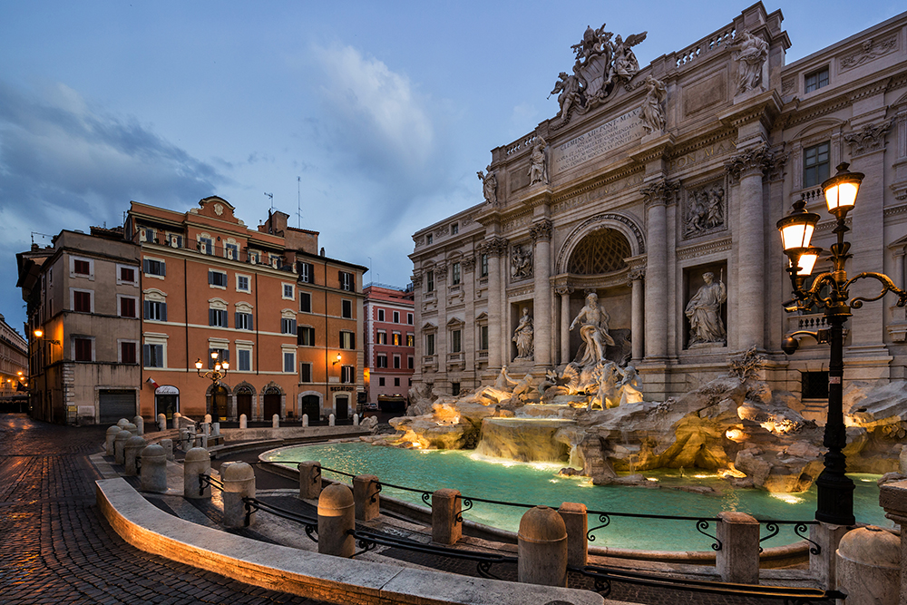 The Trevi Fountain in Rome is beautifully illuminated at dusk, its Baroque sculptures and cascading waters glowing under the soft light. Historic buildings with shuttered windows and warm-toned façades surround the square, while classic lampposts add to the romantic atmosphere.