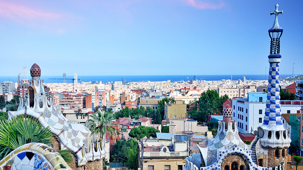 A panoramic view of Barcelona, Spain, seen from Park Güell at sunset. In the foreground, Antoni Gaudí’s iconic colorful mosaic buildings and whimsical architectural elements stand out, with vibrant blue, white, and earthy tones. The background reveals a sprawling cityscape stretching toward the Mediterranean Sea, which meets the horizon under a pastel pink and blue sky. The scene captures the blend of artistic architecture, urban life, and natural beauty that defines the city.