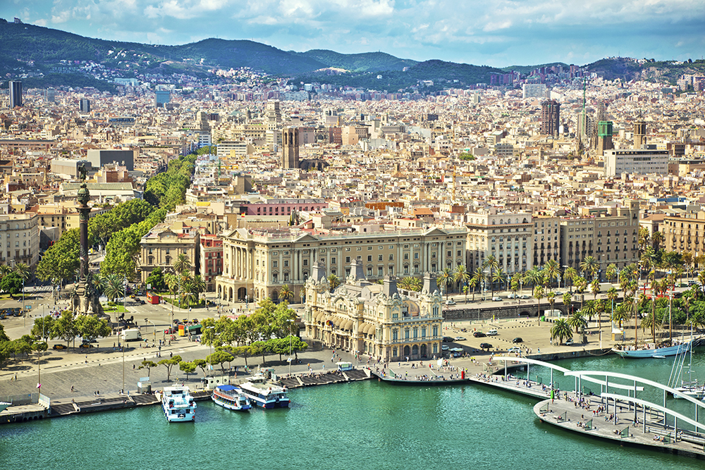 Aerial view of Barcelona’s vibrant waterfront and cityscape, showcasing the iconic Christopher Columbus Monument, palm-lined La Rambla, and the historic Port Authority building. The city stretches into the hills beyond, filled with tightly packed buildings, historic churches, and modern towers under a partly cloudy sky.