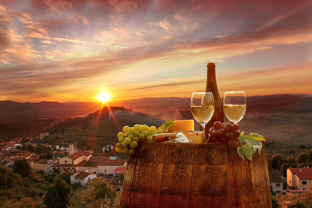 A romantic Tuscan sunset over rolling hills and a charming village. In the foreground, a rustic wine barrel holds a bottle and two glasses of white wine, surrounded by assorted cheeses, green and red grapes, and cured meats, capturing the essence of Italian countryside dining.