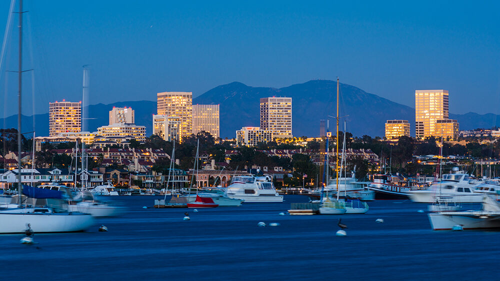 Twilight view of Newport Beach harbor with yachts and sailboats gently floating in the foreground, while the illuminated skyline of Irvine glows against the backdrop of Saddleback Mountain under a clear blue evening sky.