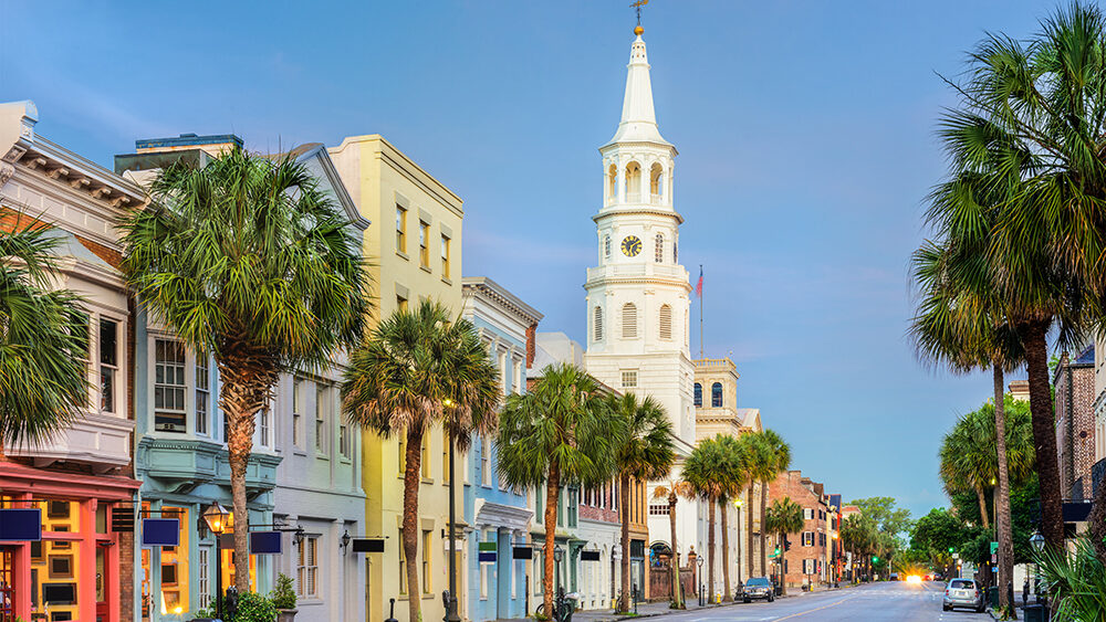 Colorful historic buildings and palm trees line a quiet street in downtown Charleston, South Carolina, with the iconic white steeple of St. Michael's Church rising in the background under a soft evening sky.