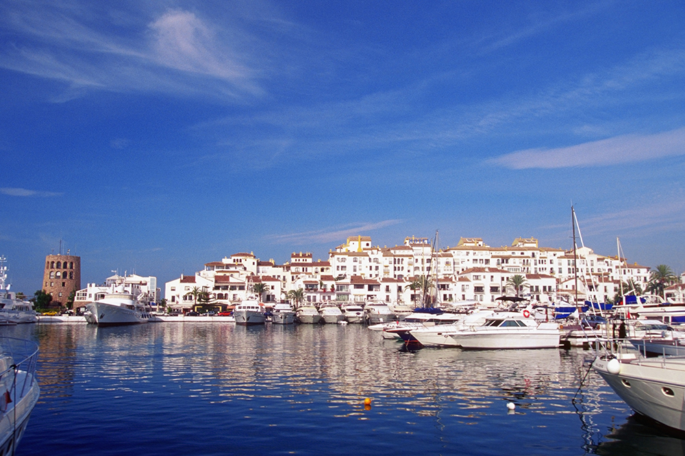 Scenic view of Puerto Banús marina in Marbella, Spain, featuring an array of white yachts docked along the calm, reflective water. Behind the boats, a cluster of whitewashed Mediterranean-style buildings with terracotta roofs rises against a vibrant blue sky. A round, stone tower stands on the far left, adding historic charm to the luxurious waterfront setting.