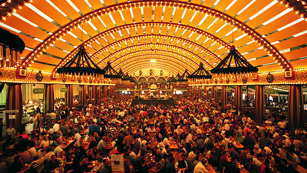 A lively and crowded beer hall at Oktoberfest in Munich, Germany, with long communal wooden tables packed with people enjoying food and drinks. The grand tent is illuminated with golden lights, and features festive chandeliers, decorative banners, and a central stage where a traditional band performs.