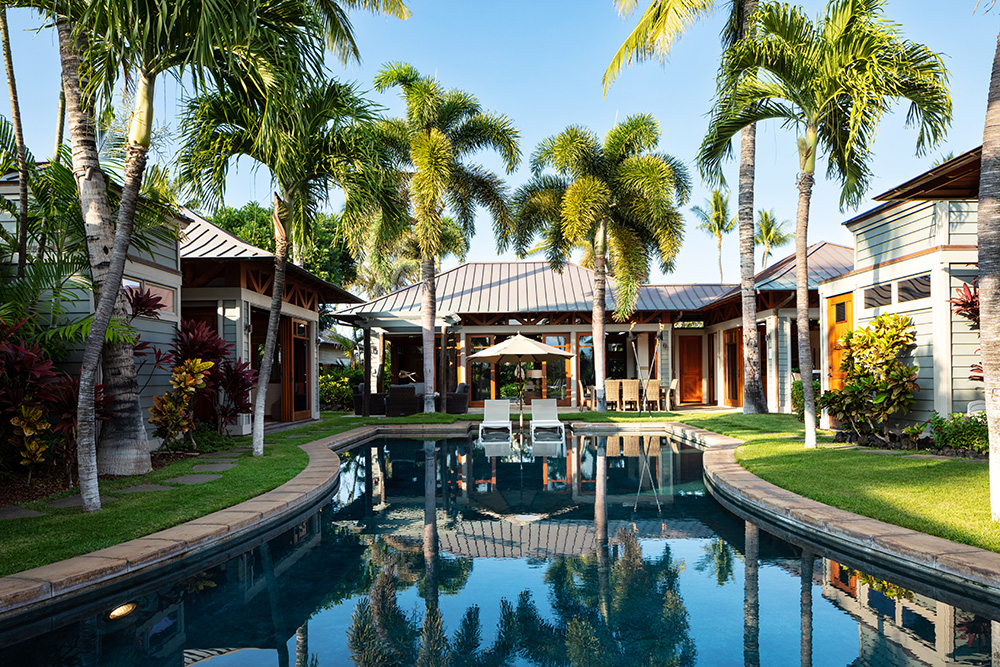 Tropical luxury villa with a lagoon-style swimming pool surrounded by palm trees, lush greenery, and multiple pavilion-style buildings with modern architecture and large glass doors.