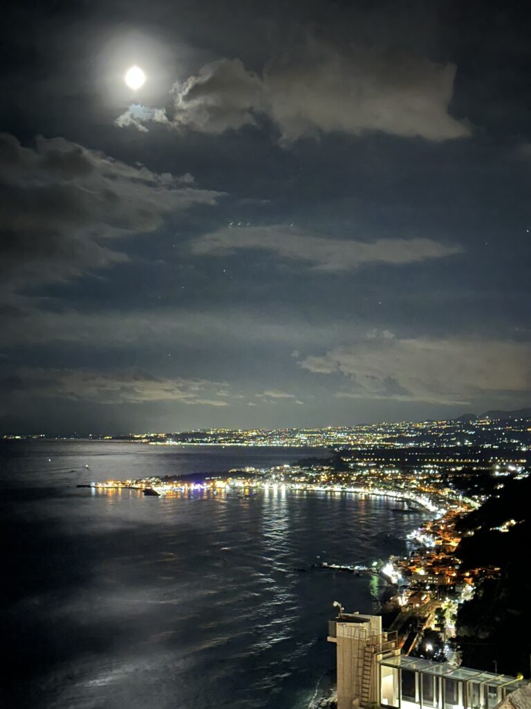 Moonlit night view overlooking the coastline of Taormina, Sicily, with city lights shimmering along the shoreline and reflections dancing on the water below.