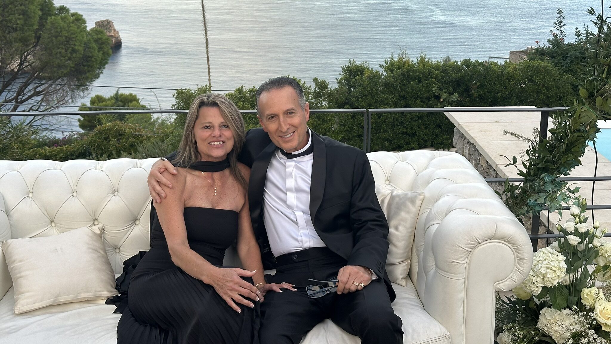 Couple dressed in formal evening attire seated on a white couch overlooking the Mediterranean Sea in Taormina, Sicily, during a special family celebration.