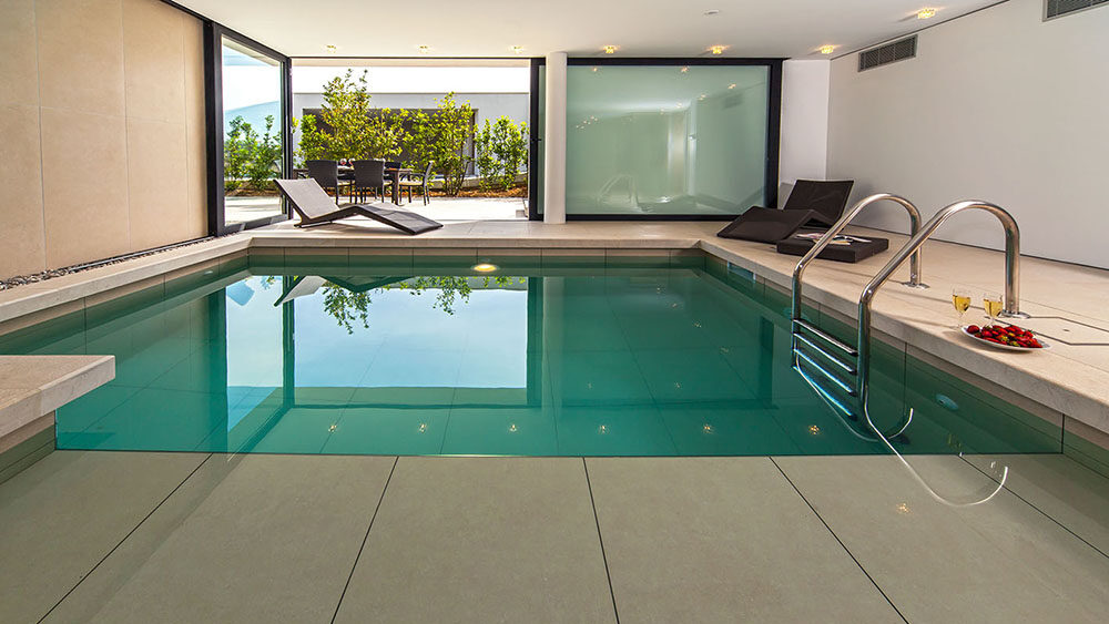 Modern indoor pool with clean tiled floors and submerged steps, surrounded by sleek lounge chairs. A tray with two glasses of white wine and a plate of strawberries rests poolside. Large sliding glass doors open to a sunlit outdoor terrace with greenery and a dining table in the background.