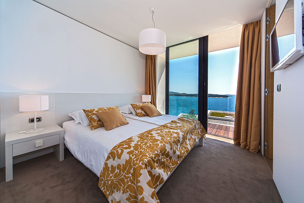 A serene modern bedroom with a large bed dressed in white linens and a golden floral-patterned throw and pillows. Floor-to-ceiling glass doors open to a balcony overlooking a stunning blue seascape. Warm golden curtains frame the view, while soft lighting from bedside lamps and a central pendant light creates a cozy, elegant atmosphere.