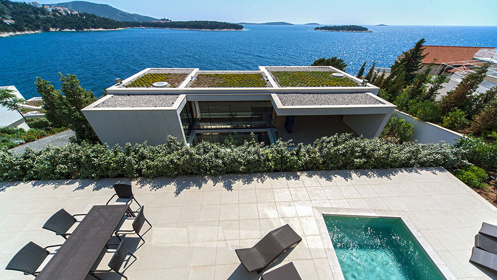 Modern terrace overlooking the Adriatic Sea with an infinity-edge plunge pool, black wicker sun loungers, and a dining table with chairs. The villa below has a flat green roof and large glass windows, while the terrace is bordered by lush greenery and offers panoramic ocean views with nearby forested islands in the distance.