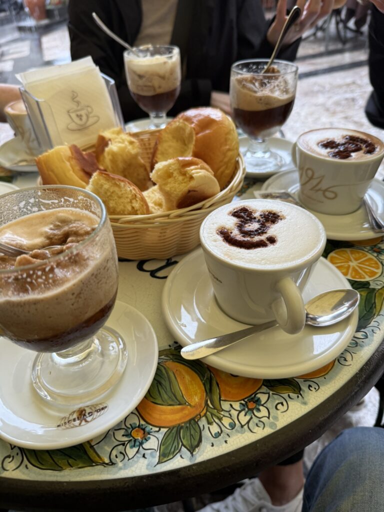 Traditional Sicilian breakfast featuring coffee granita with whipped cream, cappuccinos with chocolate art, and a basket of fresh brioche served on a colorful hand-painted ceramic table.
