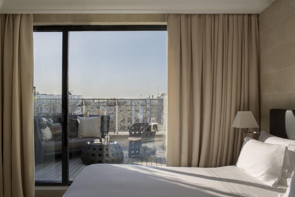 A serene view from a hotel bedroom at the Majestic Hotel in Barcelona. The image shows a neatly made bed with white linens in the foreground, next to floor-to-ceiling glass doors draped with beige curtains. Beyond the glass, a private terrace with ornate wrought-iron railings features cozy outdoor seating, including cushioned chairs and a small round table with coffee cups, all overlooking the city skyline under a clear blue sky.